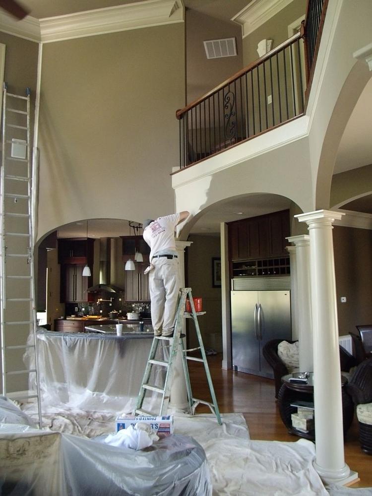 A Cawley painter on a ladder cutting in an arch in a two-story great room, with floors and furniture covered in drop cloths